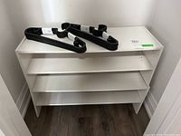 Two white laminate shoe shelves with three tiers each shown stacked in a corner with two black Ikea plastic hangers placed on top, showcasing size and surface condition