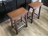Two wooden stools side by side on wood flooring near black cabinets, showing overall shape and size.