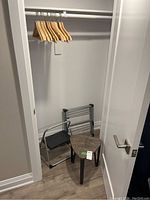 View of triangular laminate top table/stool, folding step stool, and metal/plastic laundry rack with wooden hangers in small closet space
