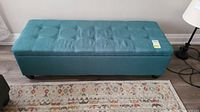 Front view of the teal upholstered storage bench with tufted top and black tapered legs on wood floor.