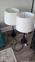 Pair of black metal table lamps with white drum lampshades placed on floor showing overall look and size.