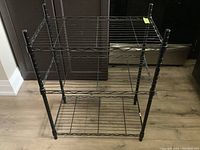 Black wire stand with three shelves viewed from a slight angle, showing the entire shelf unit and structural design against wooden flooring and dark cabinetry.