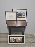 Three pieces of framed art on and in front of a dark wooden table against a wall: a larger framed print at center back, a smaller framed print at left back, and a horizontal painted wooden piece at floor front showing stylized cats.