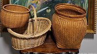 Three baskets placed on a wooden table in front of a floral painting; includes a large wicker basket, a rectangular handled wicker basket, and a round woven basket.