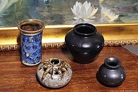 Four vases on a wooden surface against a painting background: blue pottery vase, large black vase, rustic multiple-opening vase, small matte black vase.
