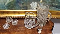 Photo showing crystal pieces on a wooden surface in front of gold framed flower painting. Includes a cut crystal jug with ice lip, vintage pressed glass pitcher, a shot glass, and two candle holders.