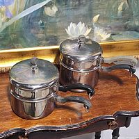 Two vintage Farberware double boiler pots with lids and black handles placed on wooden surface with flower painting in background.