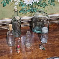 Seven vintage glass bottles arranged on a wooden surface in front of a floral painting. Includes clear and green tinted bottles of various sizes with cork stoppers, one miniature Tabasco sauce bottle, and a small bottle with a patterned surface.