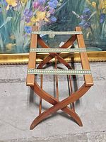 Side angle of wooden folding luggage rack showing curved cross legs, wood construction, and green leaf-patterned fabric straps.