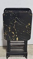 Photo showing the set of four snack tables stacked vertically folded for storage with black metal folding frames and dark green/black marble patterned tops with gold veins.