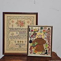 Photo showing both framed needlepoint pieces side by side on a wooden surface against a gray backdrop