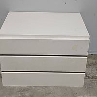 Front view of the white nightstand chest with all three drawers closed. Shows a rectangular piece of furniture with minor scuffs and marks on the surface.
