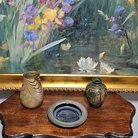 Three pottery pieces displayed on wood surface with floral painting background: gold iridescent TEFLORA vase with swirl pattern, blue pottery fish bowl with embossed fish, green etched pottery vase with turtle design.