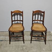Pair of wooden spindle back chairs with caned seats standing side by side against a wall on a concrete floor.