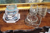 Six small glass cube-shaped dishes stacked with plastic snap lids on the left, three other glass dishes without lids on the right on a wooden surface.