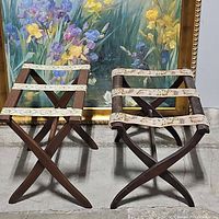 Two vintage dark wood folding luggage racks side by side with needlepoint straps, X-frame design visible