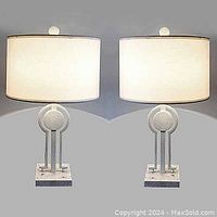 Pair of postmodern table lamps with travertine marble bases, geometric steel uprights powder coated ivory, and white drum shades with finials shown side-by-side on a gray background.
