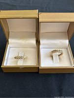 Two 14K gold rings displayed side by side in open gold-tone boxes on black surface