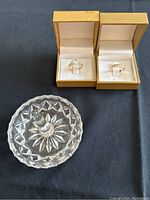 Two rings displayed in gold-tone boxes next to a glass ring holder dish on a dark surface