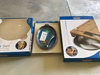 Three boxed items arranged on floor: pizza set, cast iron skillet, and cutting board set.