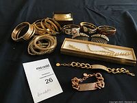 Overview photo of multiple gold toned bracelets including bangles, enameled and textured styles, rhinestone bracelets, and chain link styles arranged on black surface