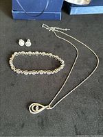 Photo showing the tennis bracelet, stud earrings, and pear-shaped pendant on the chain laid out on a black surface with blue boxes in background.