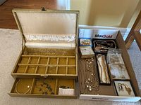 Image of vintage gold tone jewellery box open showing gold velvet compartment inserts with multiple sections and a lower tray with some necklaces inside. Next to the box is a cardboard box filled with assorted costume jewellery including chain necklaces and bracelets.