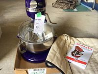 Mixer assembled with stainless steel bowl, wire whip and other attachments inside, cloth cover and manual beside it.