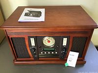 Full front view of the wooden music center showing its wood finish, speaker grills, control panel, and instruction booklet on top.