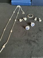 Sterling silver necklace on left with spaced white stones, several pairs of stud earrings and a ring on right, all on black surface