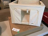 Beige Organizeme collapsible fabric storage tote placed on its cardboard box, showing zippered front and fabric material in light cream/beige color.