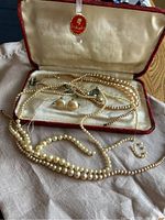 Top view of multiple cream pearl-like bead strands with clasps and a pair of clip-on earrings in a fabric-lined red case