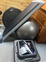Photo showing black exercise step platform angled on top of a silver-gray yoga ball, with the compartment on the platform filled with workout accessories like resistance bands, water bottle, and straps.