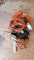 Photo showing several orange extension cords, a black cord, a white power strip, and a handheld trouble light with protective metal cage, all laid on a wooden floor.