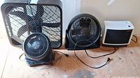 Group photo showing one large Pelonis box fan, two smaller Honeywell table fans, and one Thermaflo Seabreeze heater on a wooden floor surface.