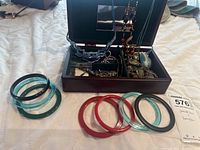 Overall view of the open jewelry box with necklaces and bracelets inside; several colorful bangle bracelets placed outside the box on a white surface.