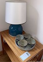 Photo showing lamp with blue ceramic base and white fabric shade, placed on wooden side table.