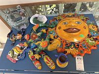 Photo showing full view of multiple artisan hand painted Mexican pottery and wall decor items arranged on a table, including sun face plaque, lizards, fish plate, jug, green pottery piece, and bird figurine.
