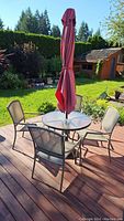 Full patio set on wooden deck with green grass and garden background, showing the table, four chairs and closed red umbrella.