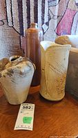 Photo showing three vintage stoneware items: tall brown bottle, beige pitcher with carved patterns, Kevin Head signed pottery vase with chips.