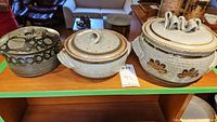 Three pottery crocks displayed on wooden shelf, each with a lid and distinct glaze and decoration.