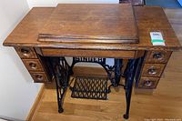 Front view showing the oak wood table with six drawers and black cast iron treadle with 'SINGER' logo.
