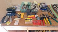Full view of table with assorted hand tools: staplers, boxes of staples, hand snippers, measuring tape, ratchets, sockets, multimeter box, and other tools.