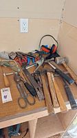 Photo showing an assortment of hand tools including hammers, pliers, scissors, wrenches, saw, and wire cutters on a wooden surface.
