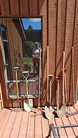 Photo showing six long-handled yard tools consisting of three shovels with red D-shaped grips, one narrow spade, a garden fork, and a rake leaning against a wall with wooden plank siding.