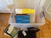 Plastic container holding various painting surfaces including canvases and paper, assorted brushes and a zippered art tote on the floor in front of the container