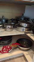 Various stainless steel pots with metal lids, non-stick frying pans with red and wooden handles on shelves.