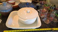 Various ceramic casserole dishes, white ceramic dish with lid and knob handle, Pyrex glass measuring cups and jugs on counter with yellow measuring tape for scale.