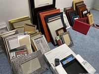 Multiple picture frames arranged on floor including metallic silver, wood-tone, and decorative styles, plus boxed digital photo frame and power adapter visible.