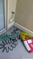 Photo showing three coiled garden hoses connected to an outdoor spigot, one in yellow contained in branded storage bag, two green with spray attachments.